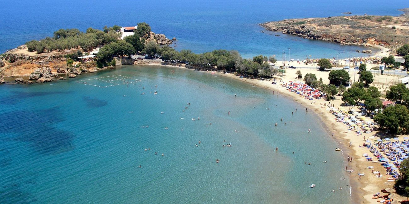 Glaros beach with shallow blue waters and sandy shore and the little peninsula with saint Apostoles church surrountded by greenery